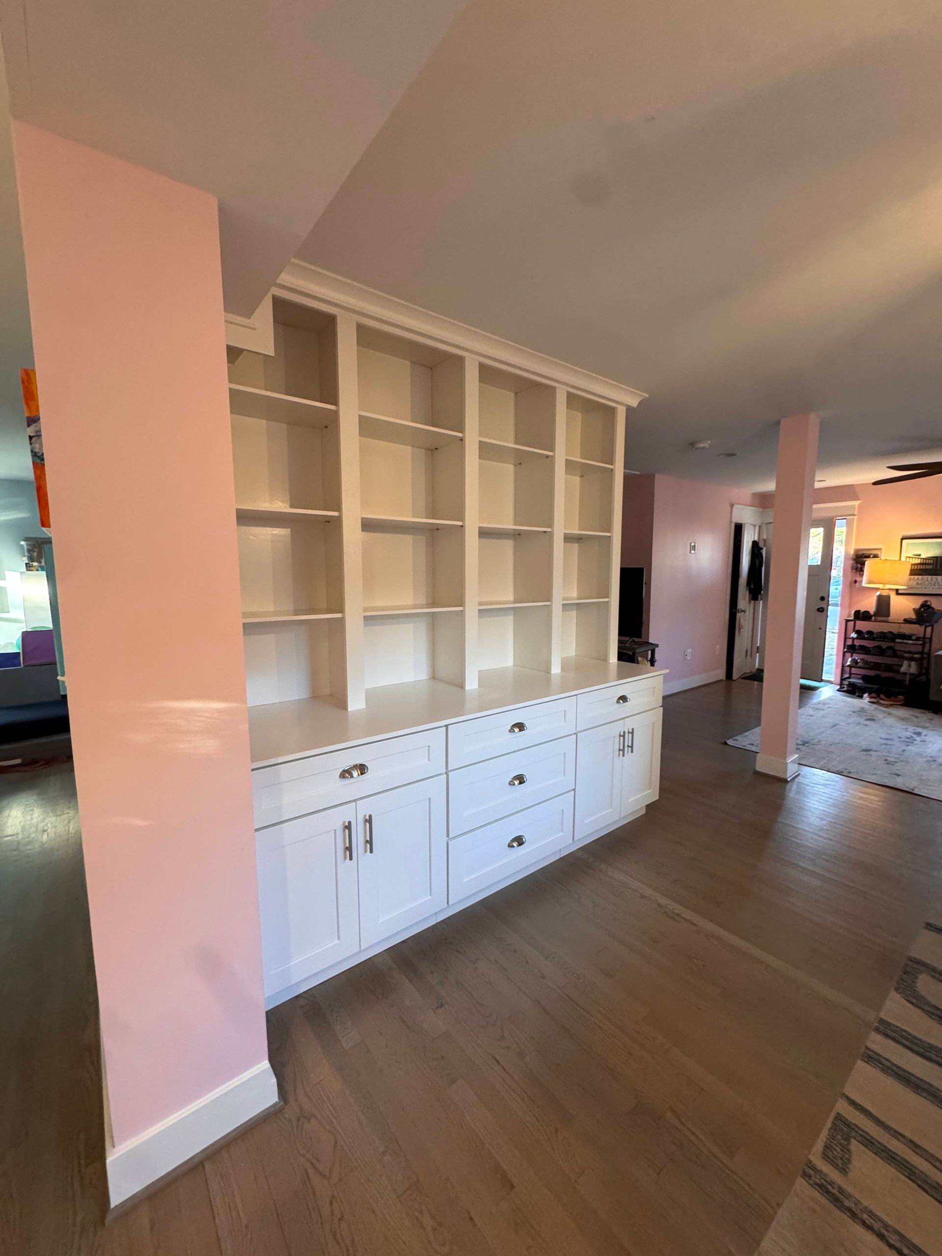 White built-in shelves and cabinets dividing two rooms. Pink walls, wood floors.