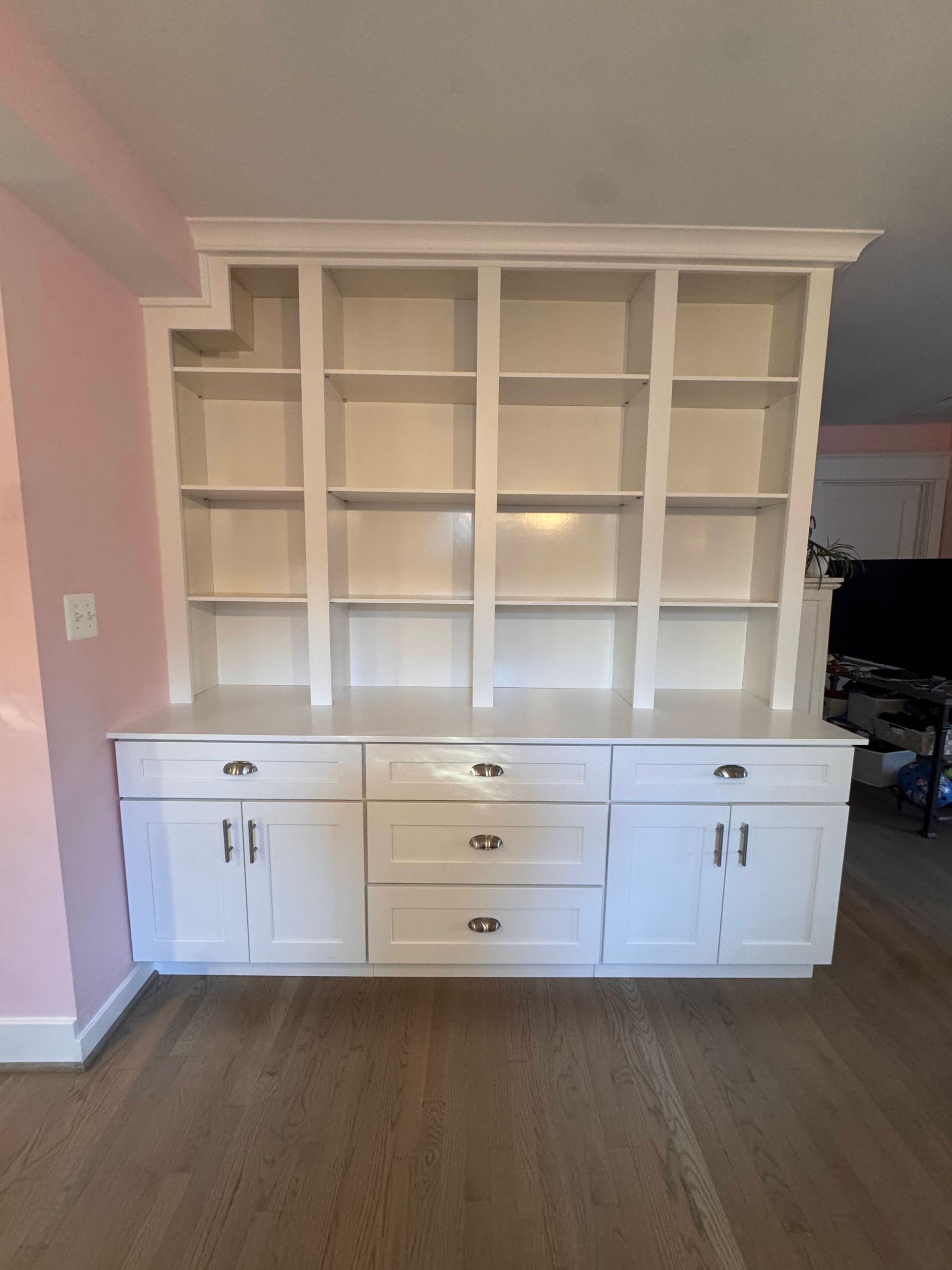 White built-in cabinet with bookshelves and drawers against a pink wall and wood floor.