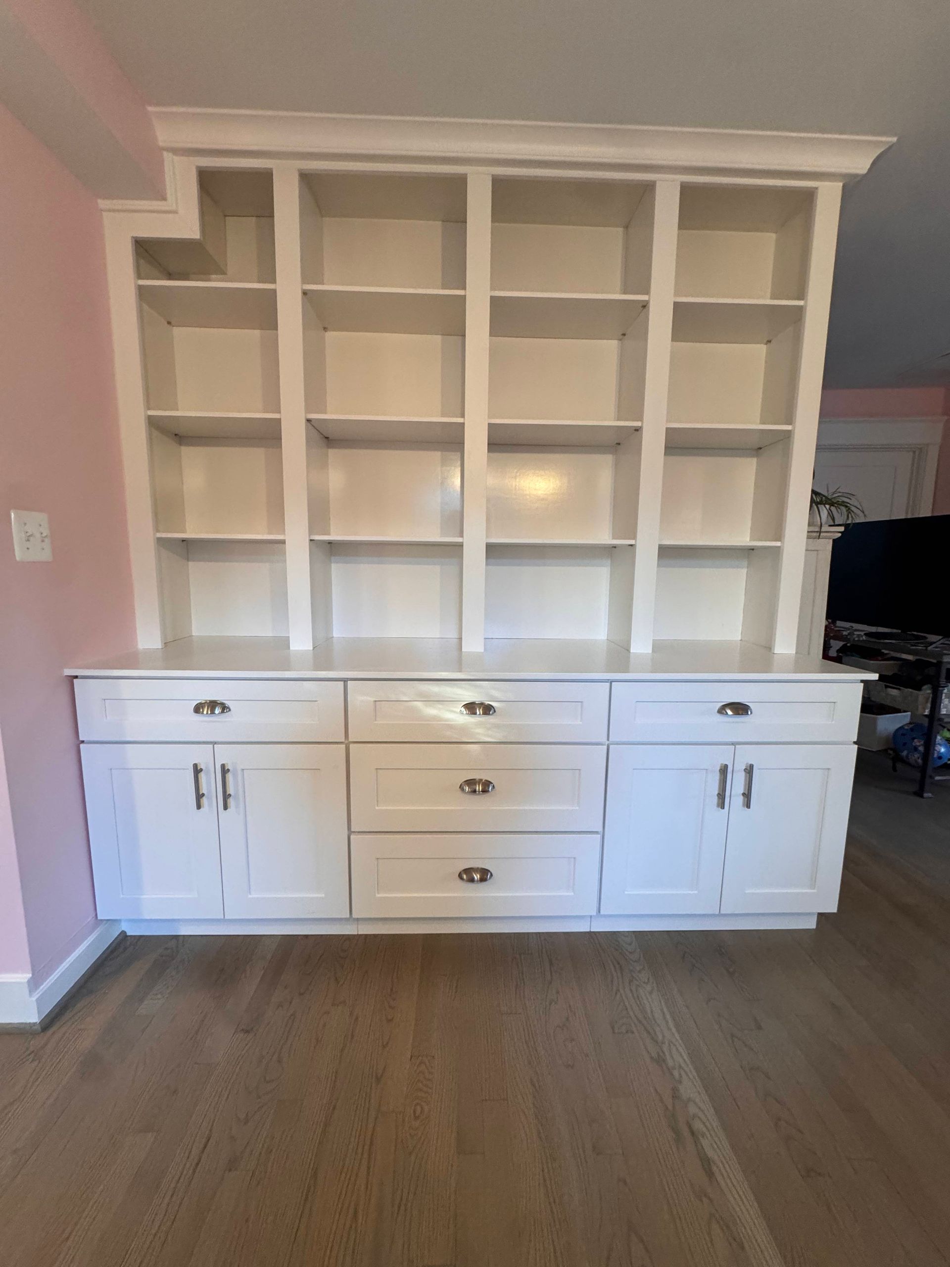 White built-in cabinet with open shelves above and doors/drawers below; wood floor and pink wall.