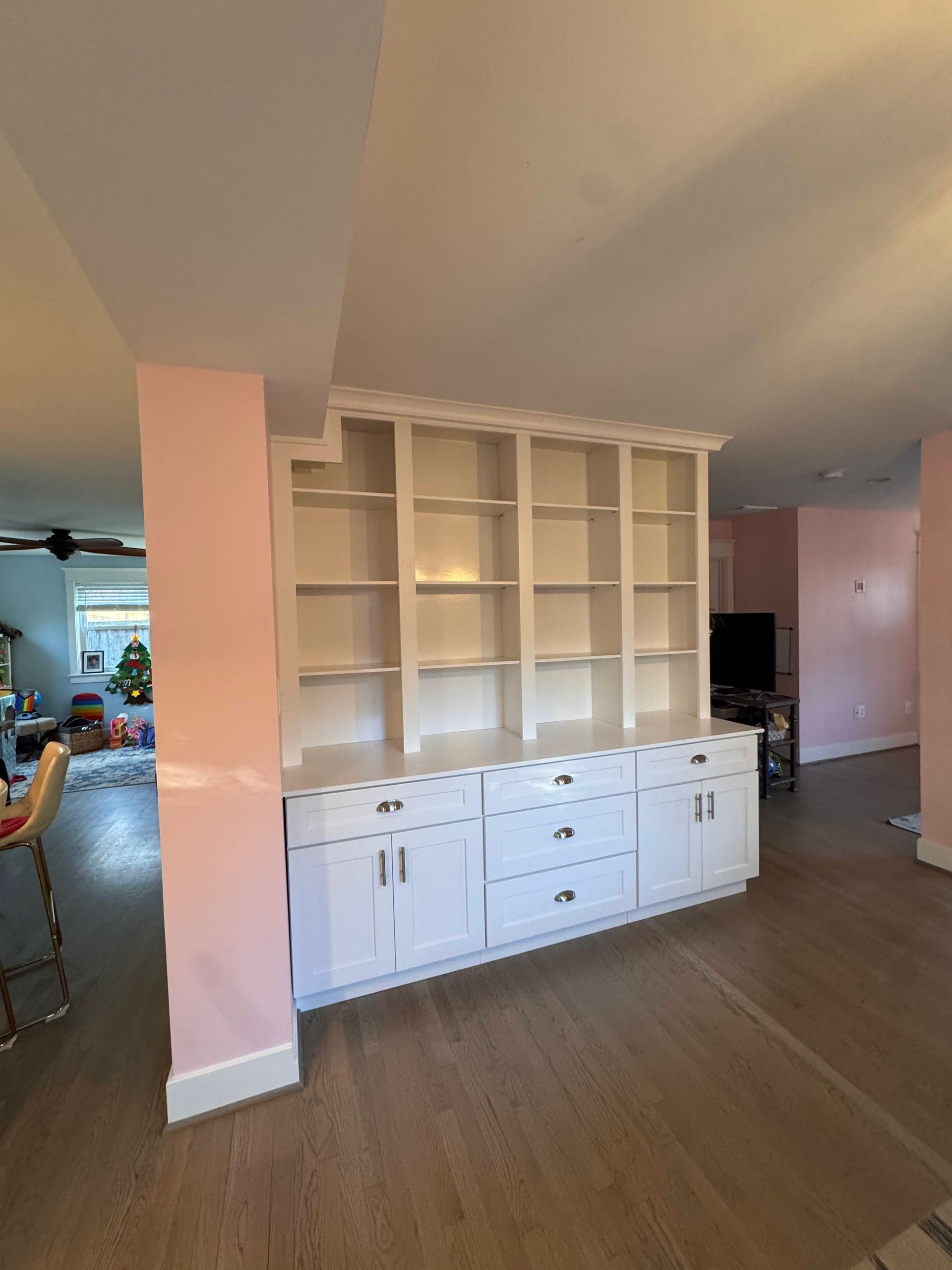 White built-in shelving unit with cabinets, centered on a light wooden floor, adjacent to a pink wall and doorway.