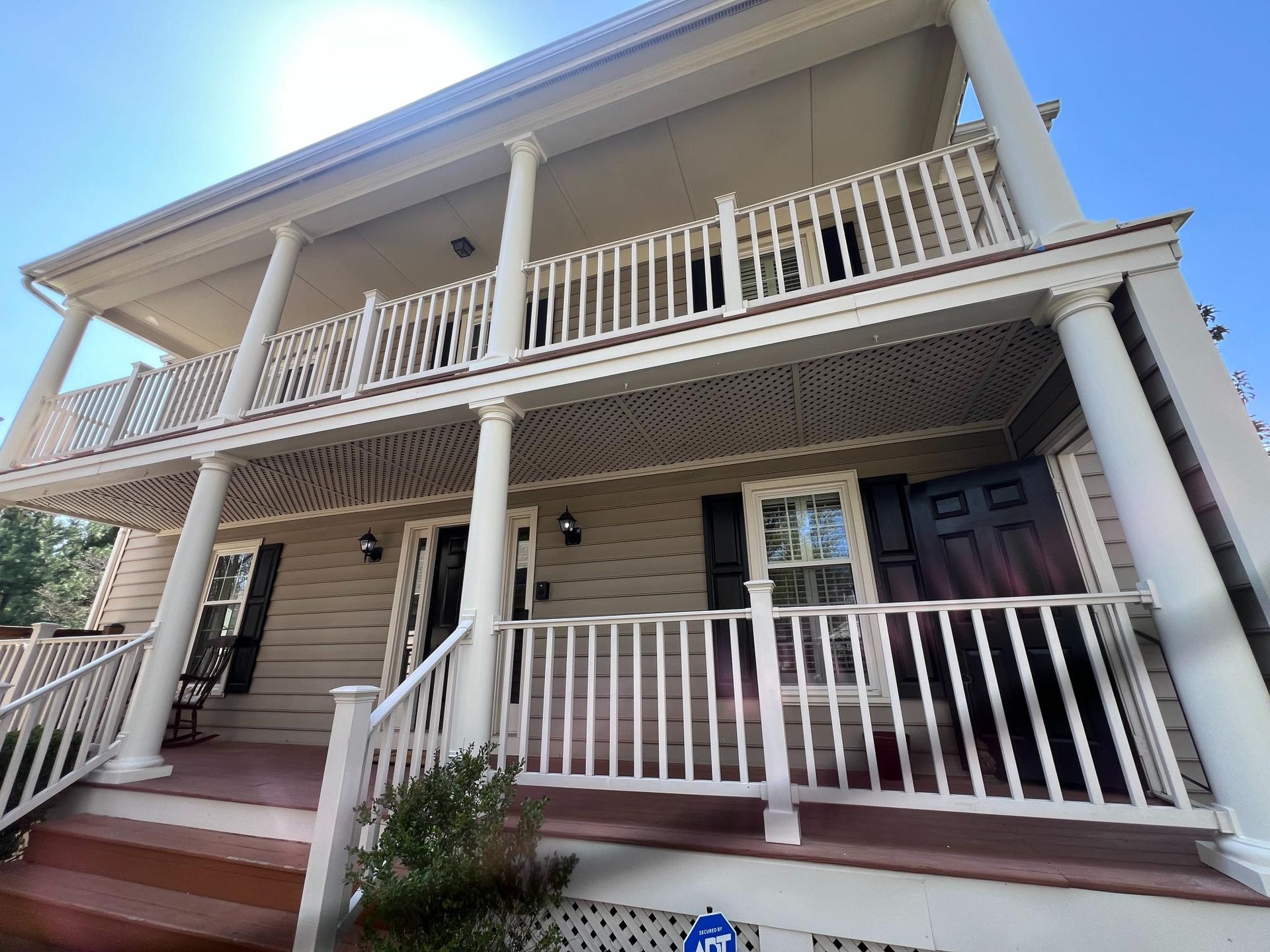 A two-story house with a wooden porch and balcony, white columns, beige siding, and a dark front door.