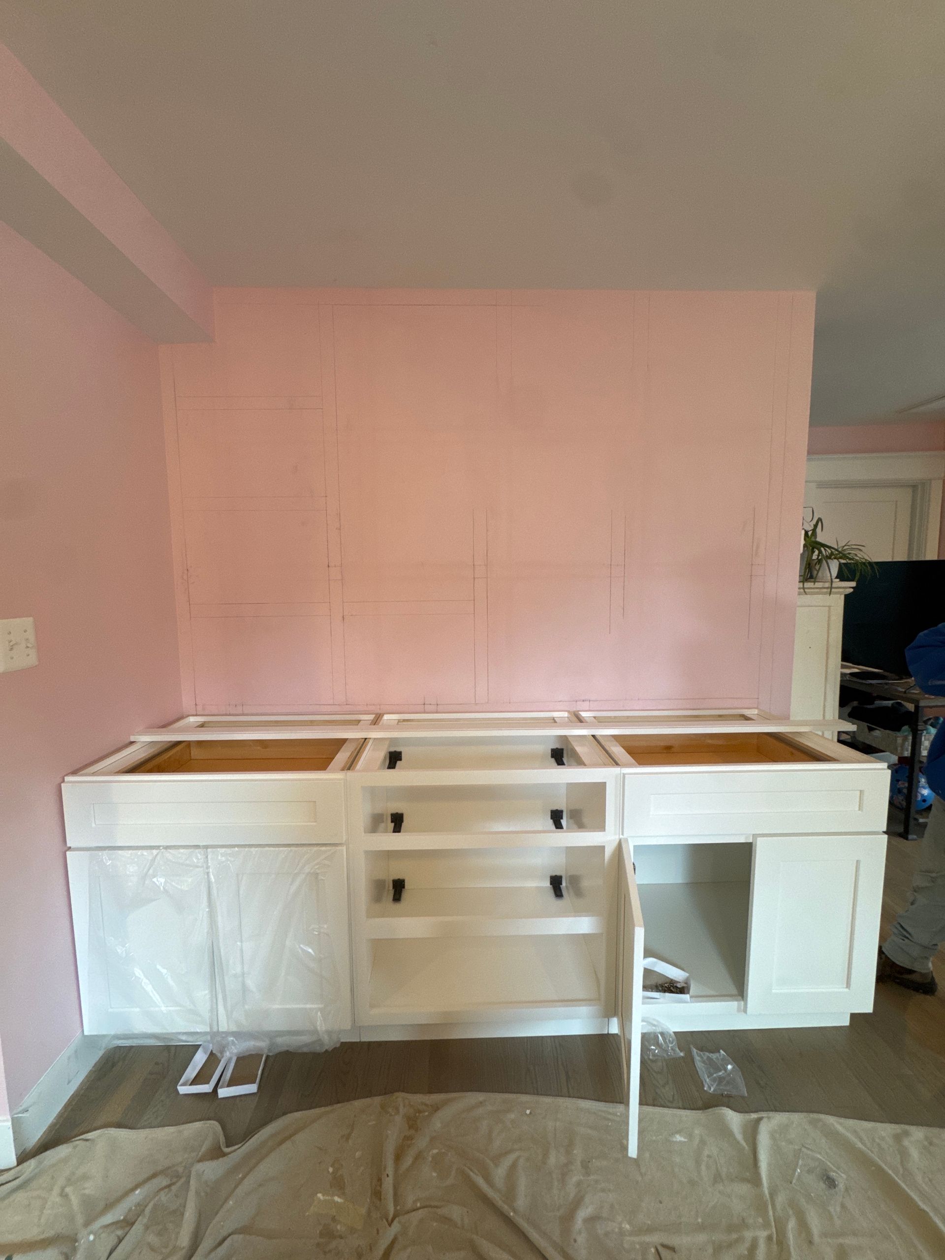White kitchen cabinets installed against a pink wall, undergoing construction.