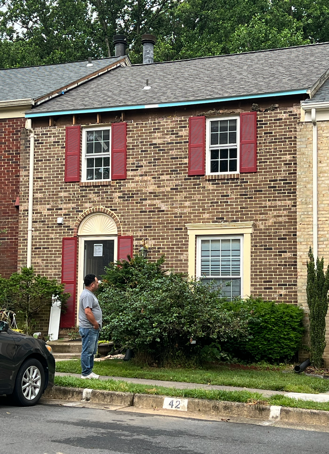 A man is standing in front of a brick house with red shutters.