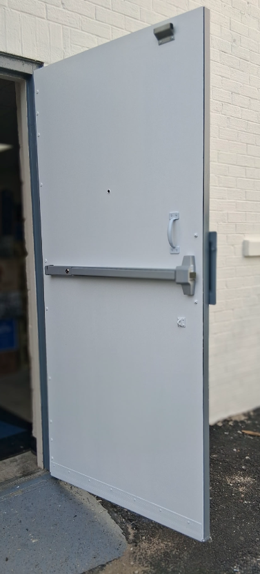 A white metal door ajar. The door has a lever, handle, and a silver crash bar. The door is set in a white brick wall.