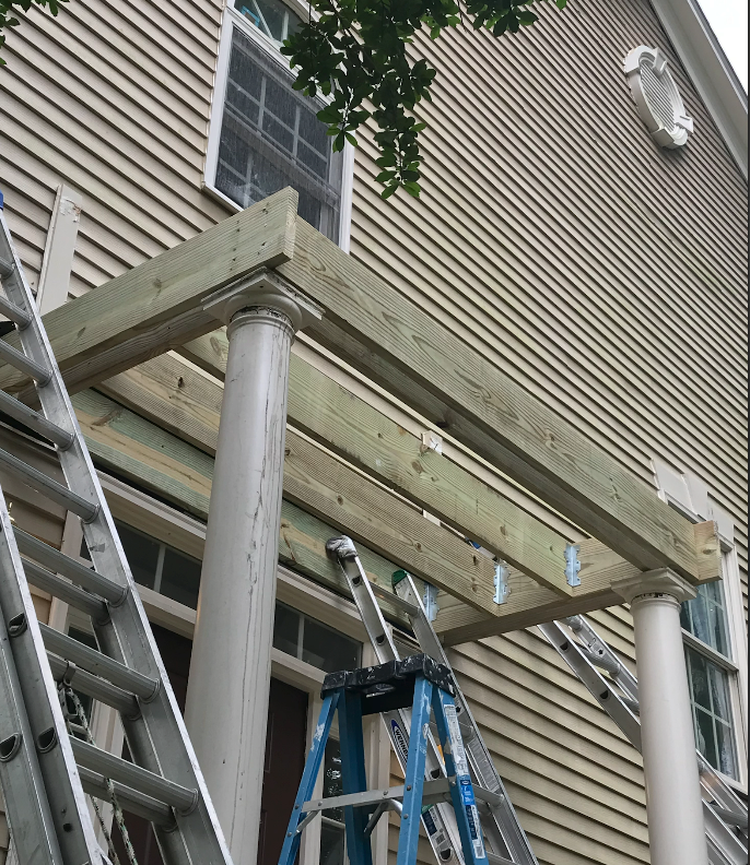 Construction of a porch roof in progress; lumber frame supported by white columns, with ladders and a blue stepladder present.