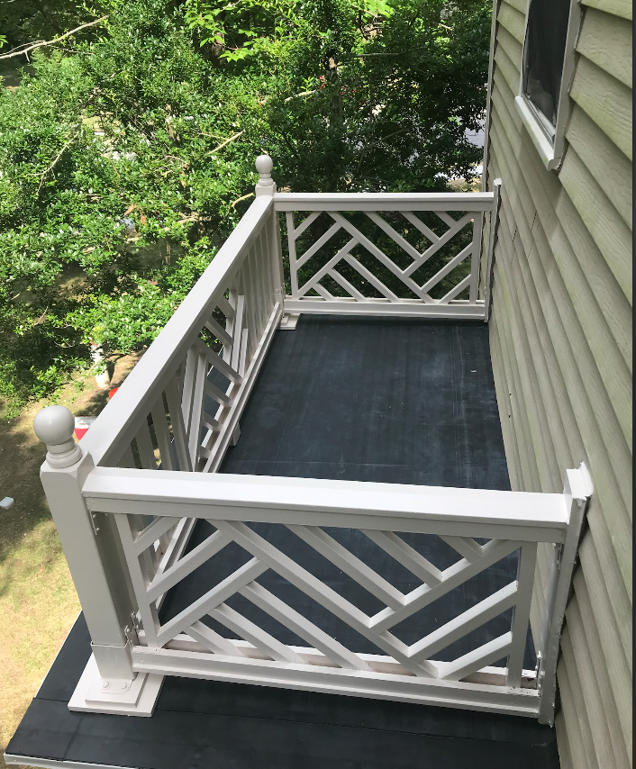 White wooden balcony with a patterned railing attached to a beige building. The balcony floor is covered in black material.