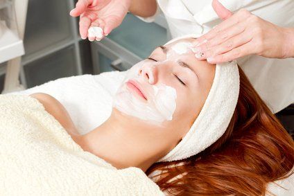 A person relaxing on a spa bed while an aesthetician applies a white face mask.