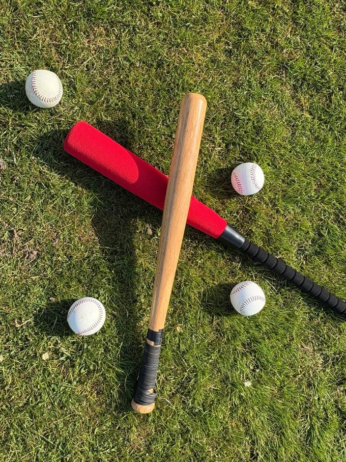 Two baseball bats, one red, one wooden, cross on grass with four baseballs.