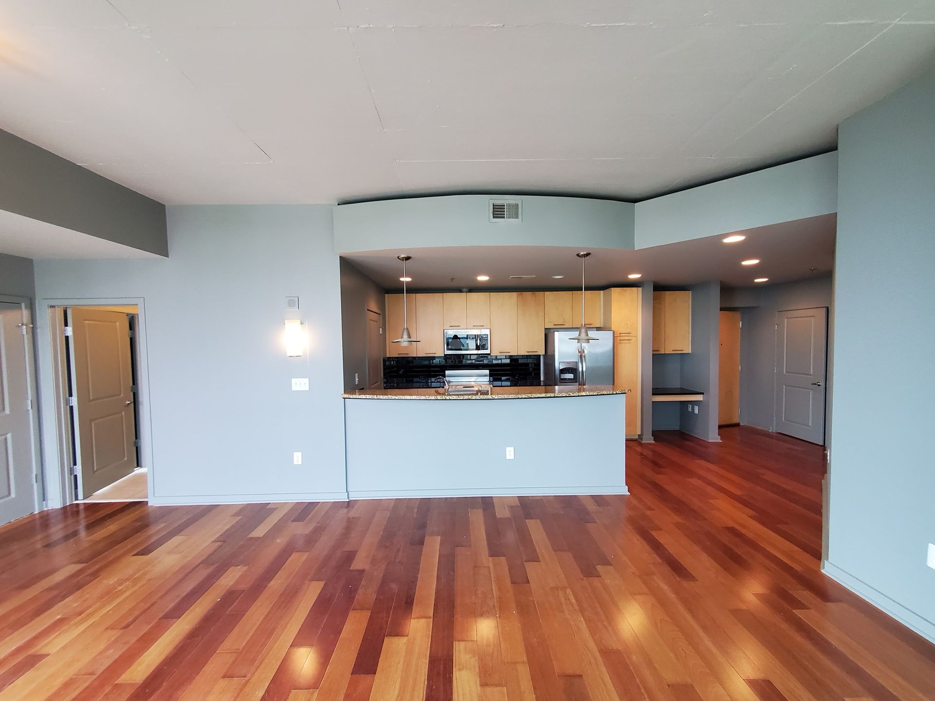 Spacious condo interior with hardwood floors, light blue walls, and a view into the kitchen area.