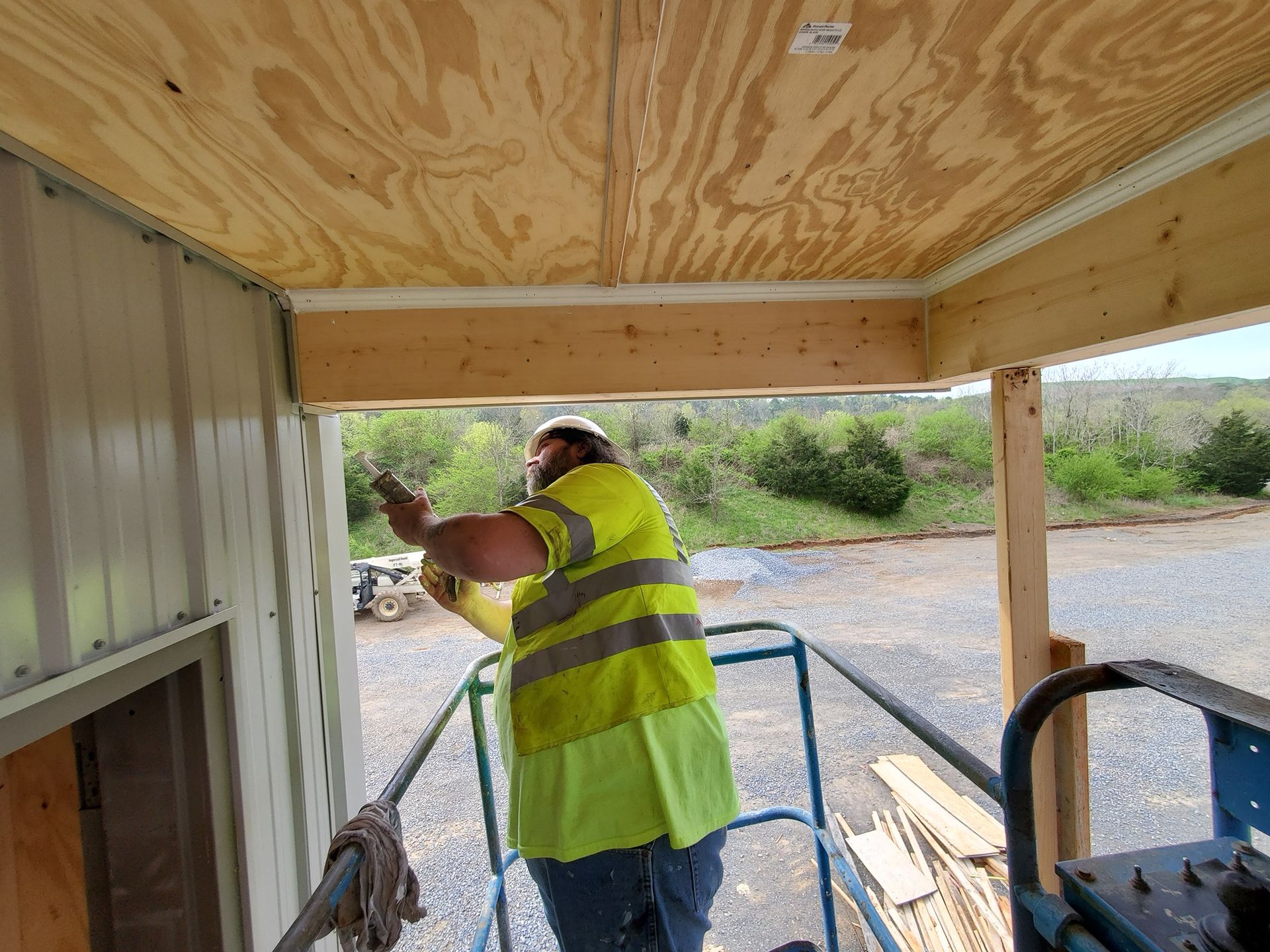 Construction worker in a safety vest on a lift installing trim under a wooden overhang next to a metal building.