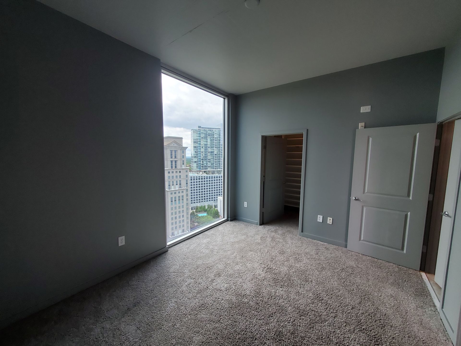 Empty room with large window, gray walls, and carpet. Door and closet visible. City view.