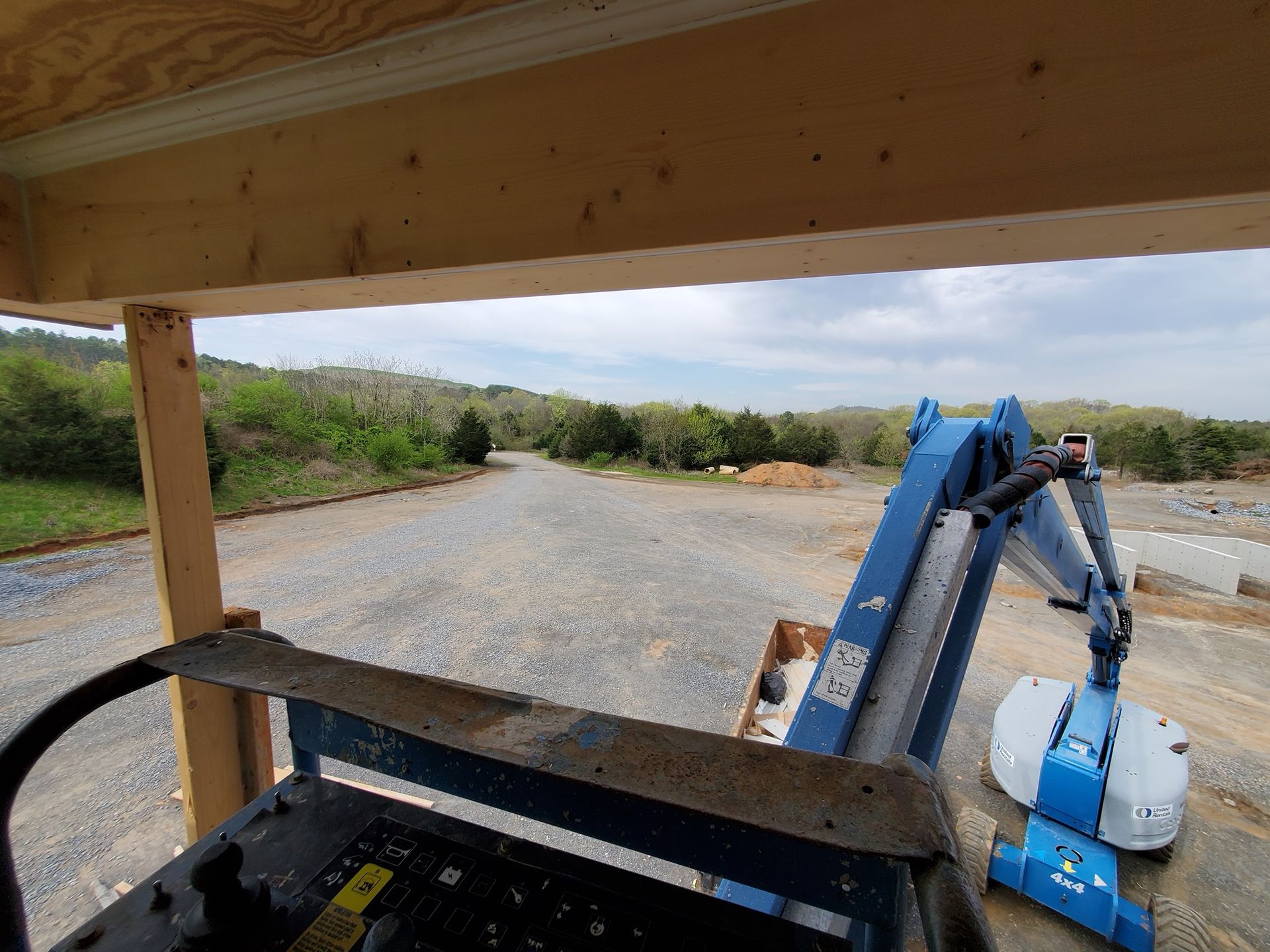 View from a construction lift overlooking a gravel road leading to a treeline. Blue machinery visible.