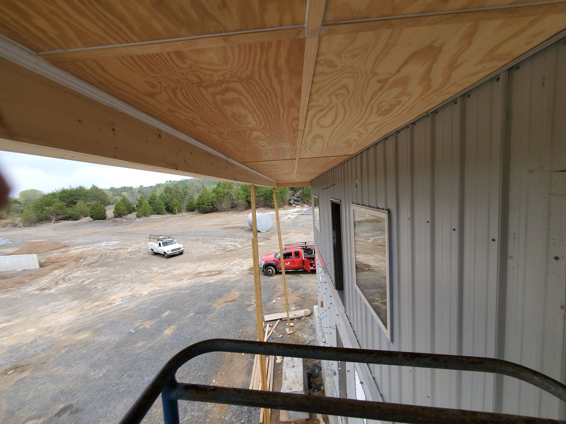 Exterior view of building under construction with metal siding and a plywood overhang, vehicles in the background.