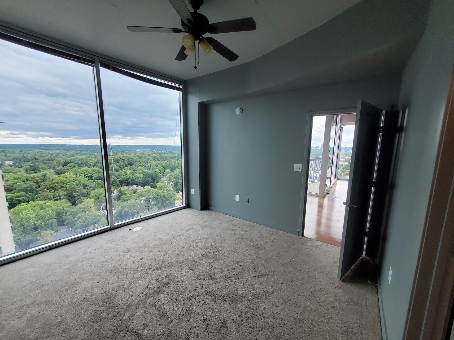 Empty room with large windows overlooking a green landscape. Carpeted floor, blue-gray walls, open doorway.