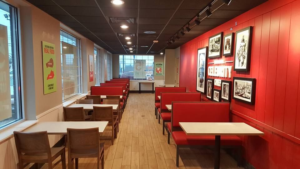 Restaurant interior with booths, tables, and framed artwork on a red wall. Brown chairs and a black ceiling.