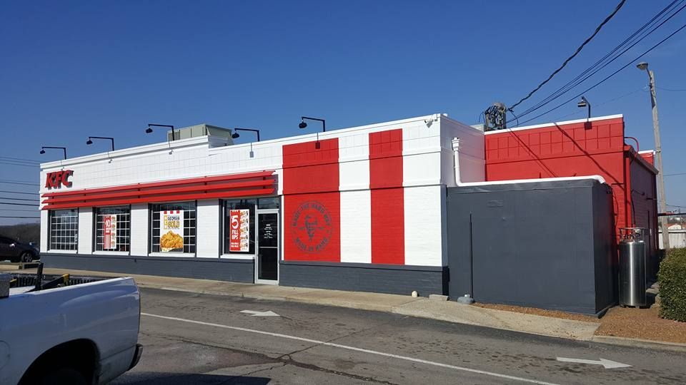 Exterior of a KFC restaurant; white and red building with a drive-through lane under a blue sky.