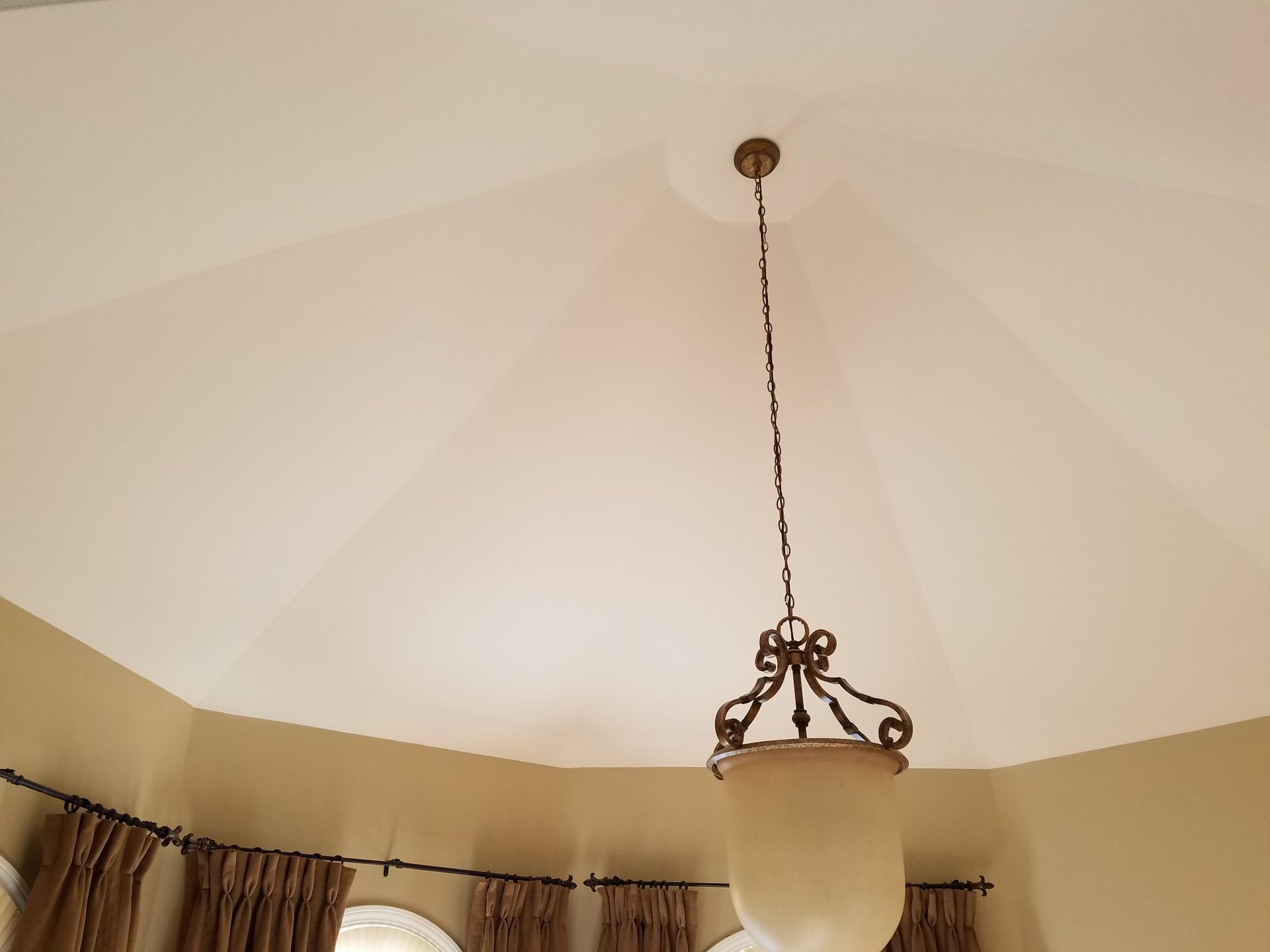 A chandelier hangs from the center of a white, octagonal ceiling in a room with tan walls and curtains.