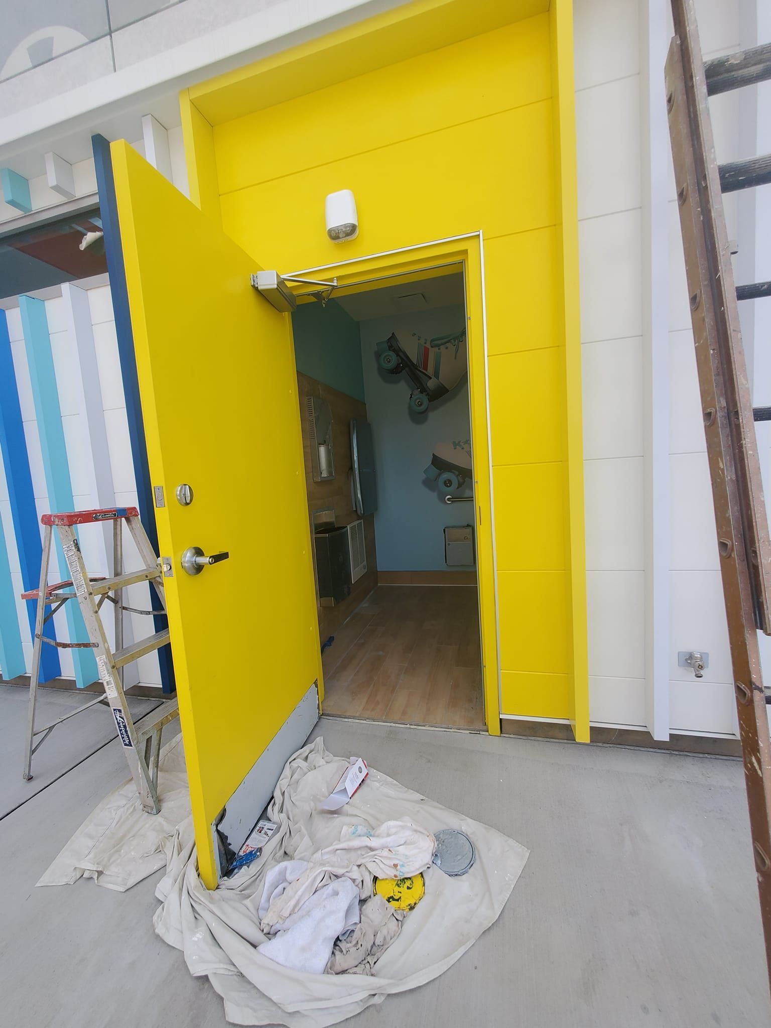 Yellow door, partially open. A small stepladder sits to the left. A paintbrush and cloth lie on the floor.