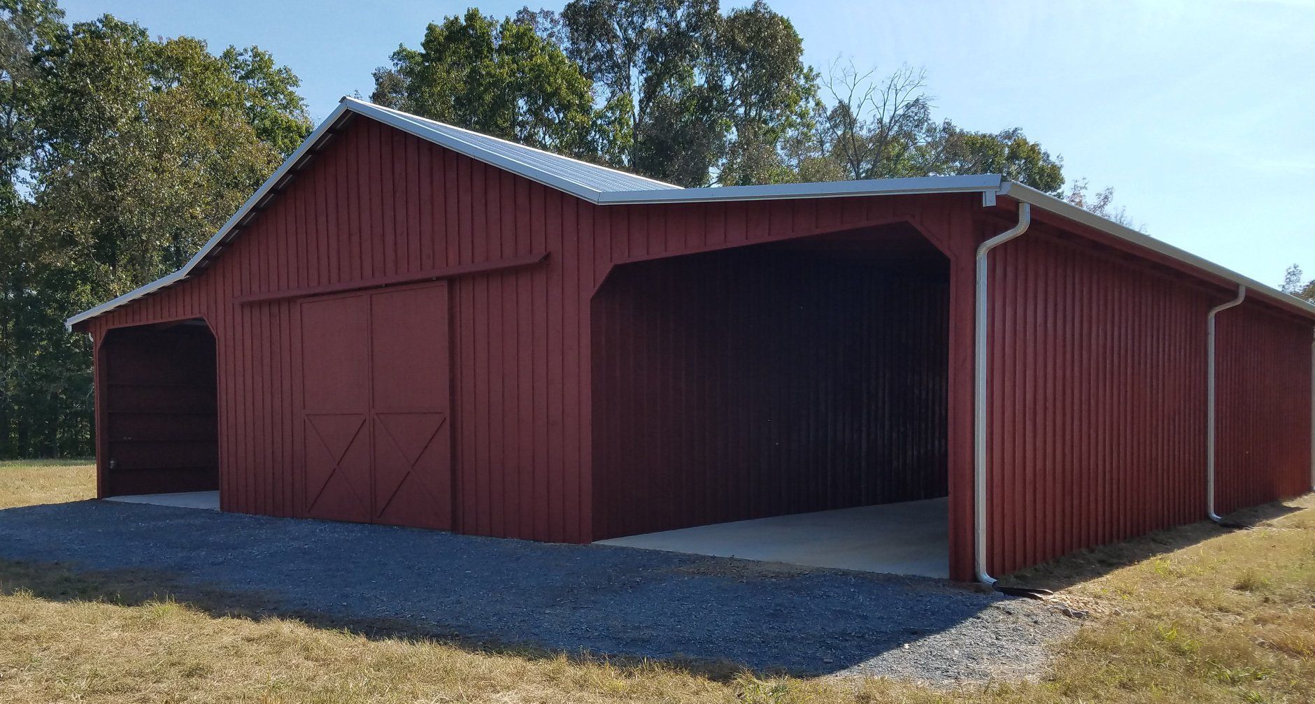 Red barn with a metal roof and open-sided carport on a gravel base, set in a grassy area with trees.