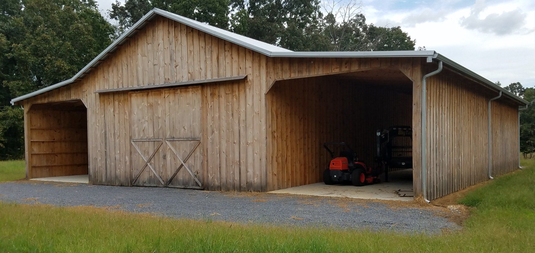 Wooden barn with open bays, gravel driveway, and a tractor inside. Green grass and trees in the background.