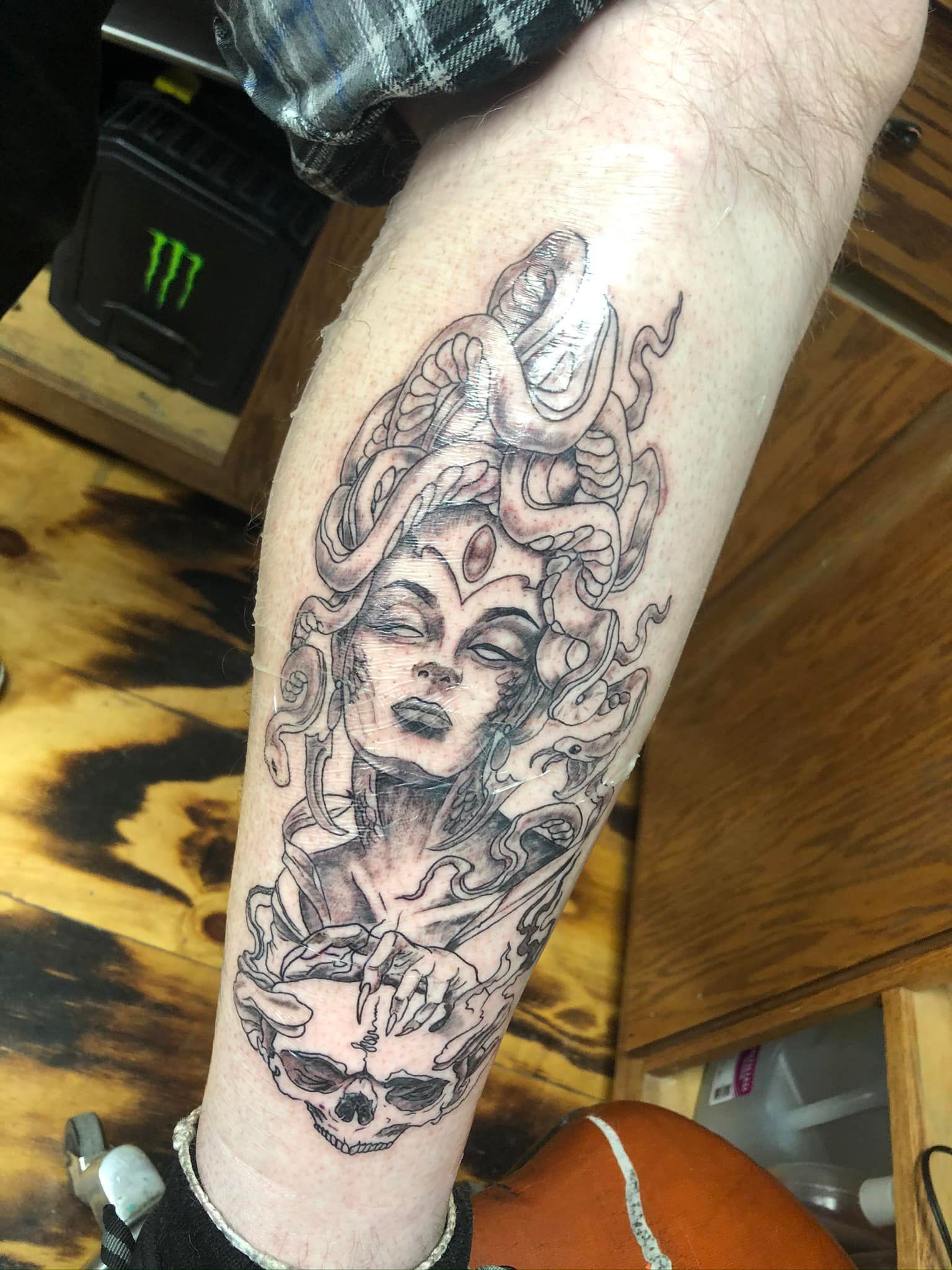 Tattoo of a woman with snakes for hair and a skull, on a person's leg. Black and white.