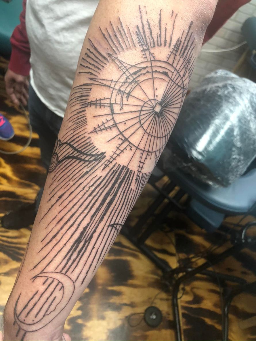 Forearm tattoo: radiating sunburst with lines, compass-like center, crescent moon, and clouds.