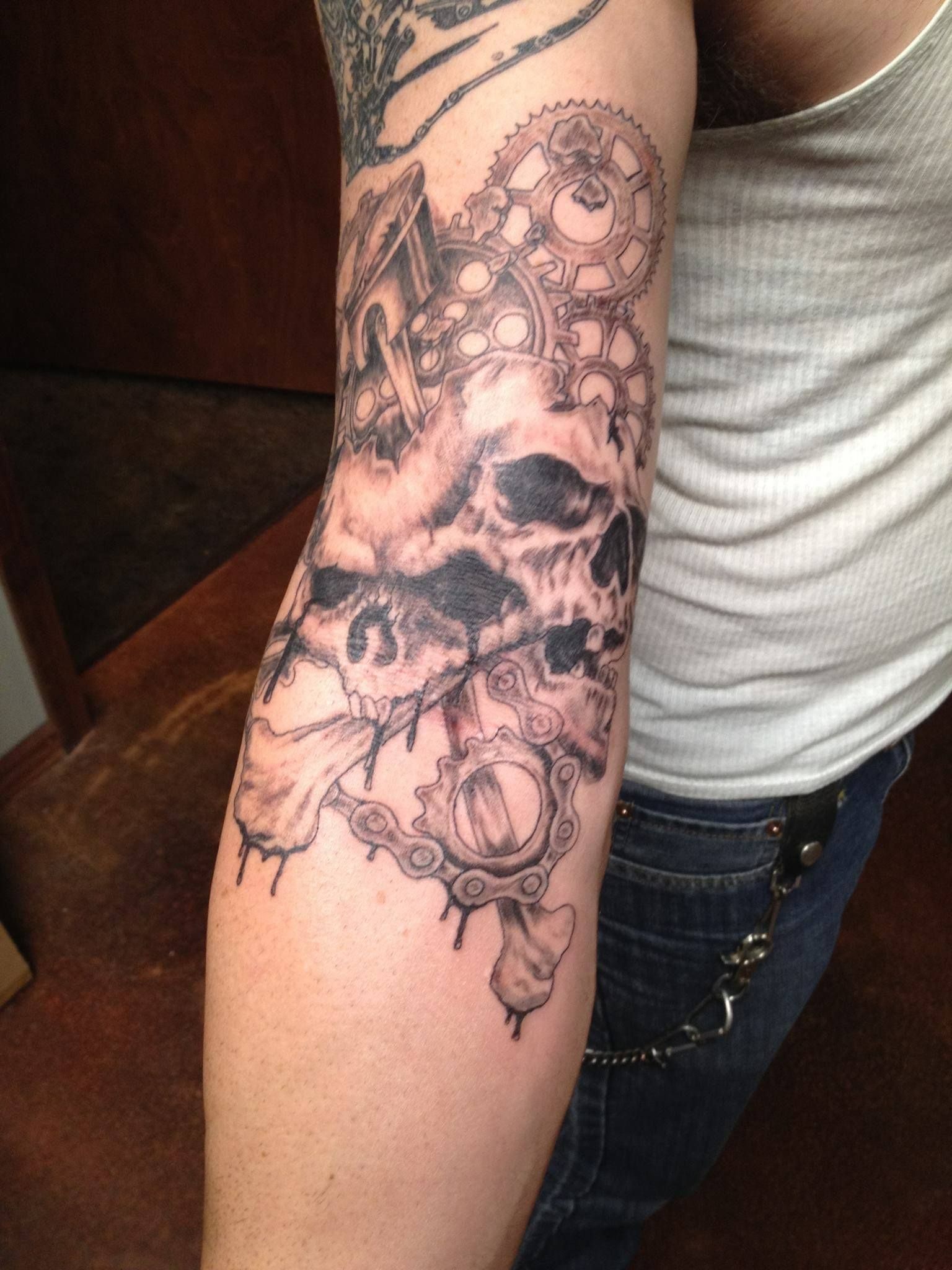 Black and gray forearm tattoo: skull with gears and clockwork elements. Person wears a white tank top and jeans.
