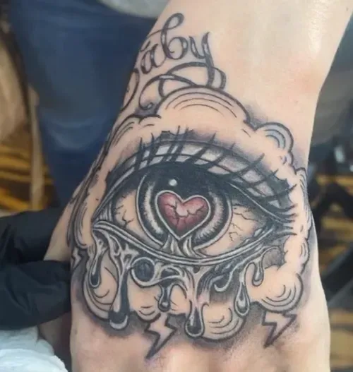 Hand tattoo: eye with heart-shaped pupil, tears, clouds.
