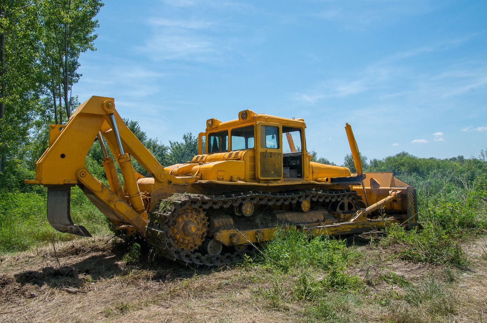 Yellow bulldozer with a saw attachment on a grassy field under a blue sky.