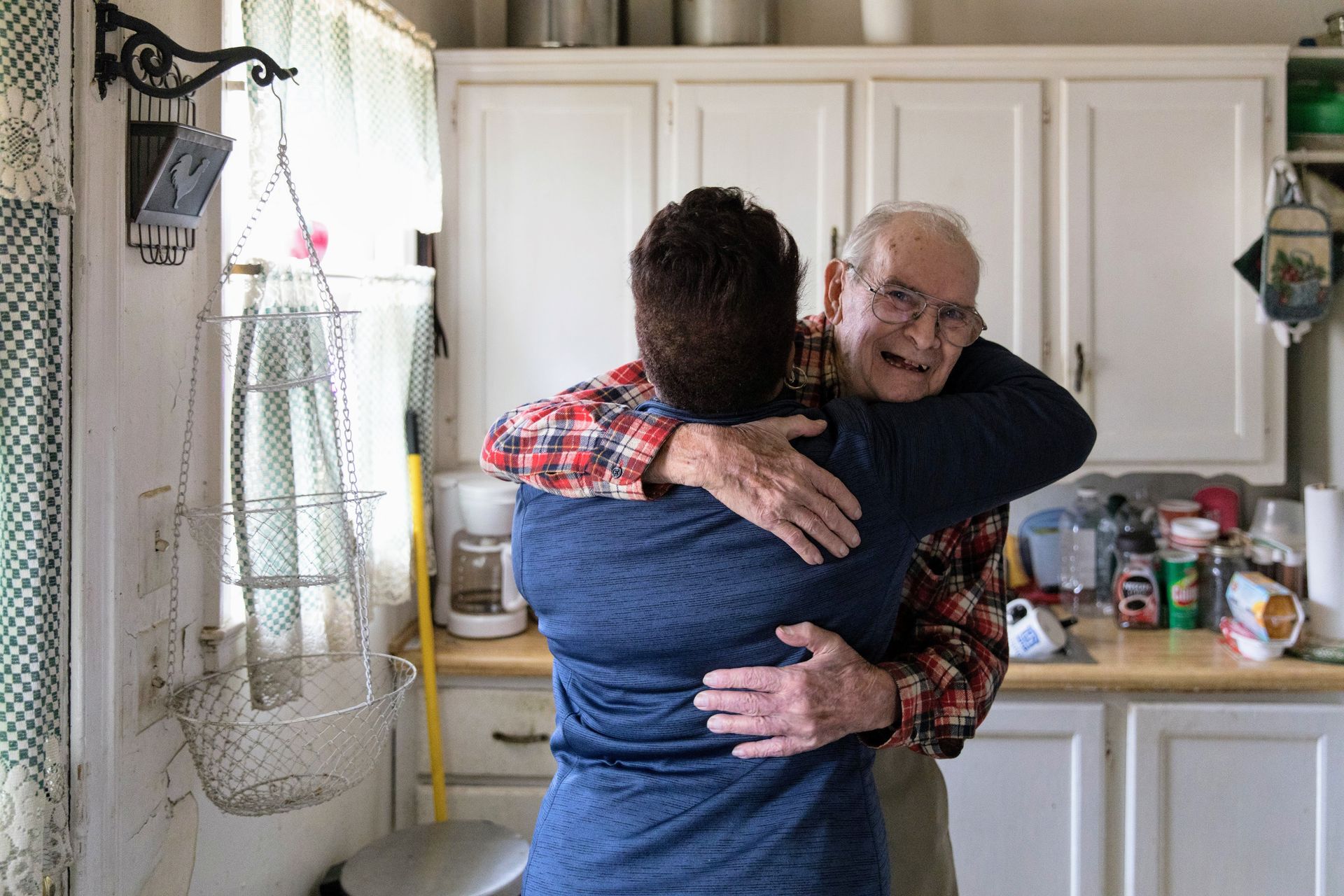 An elderly man is hugging a younger man in a kitchen.