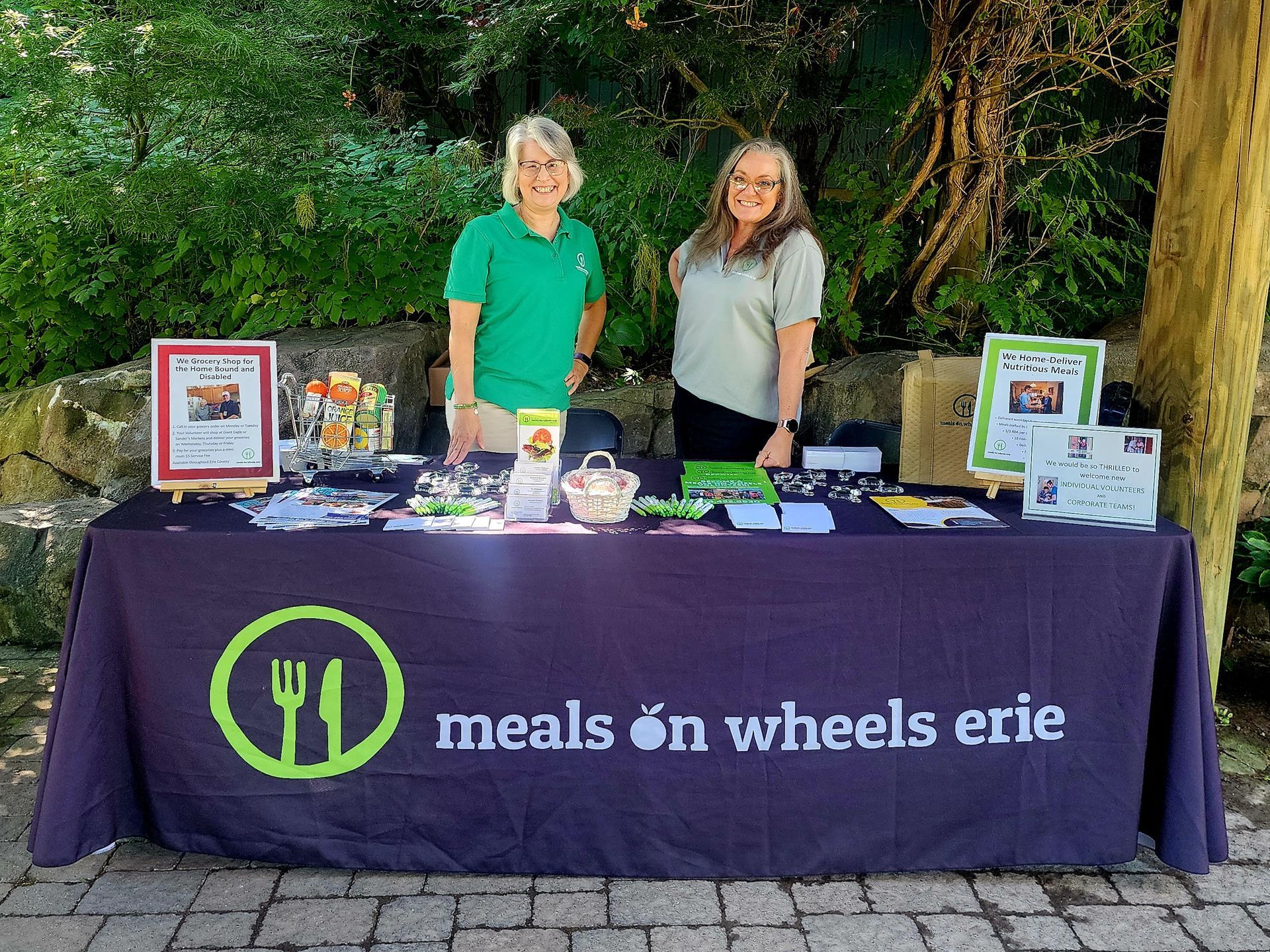 Two women stand behind a table that says meals on wheels erie