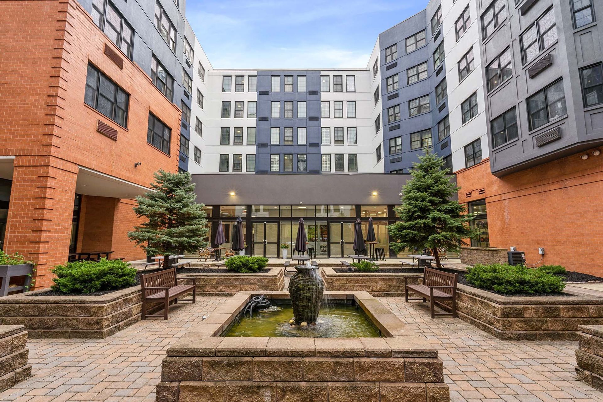 Outdoor communal courtyard with a central fountain, planters, and benches surrounded by modern apartment buildings Parc at Lyndhurst in Lyndhurst, NJ.