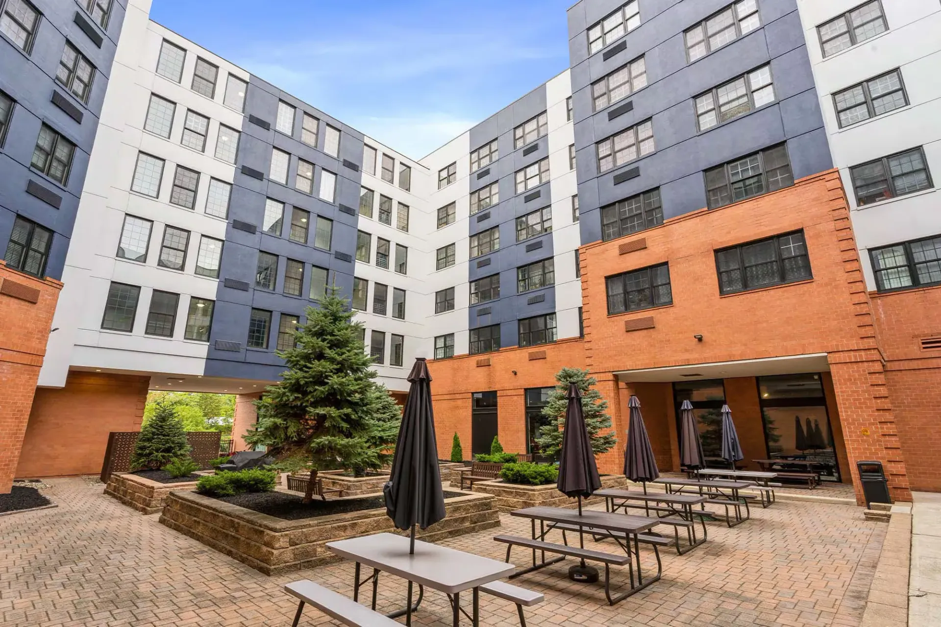 Outdoor community courtyard with brick buildings, trees, planters, and picnic tables with umbrellas Parc at Lyndhurst in Lyndhurst, NJ.