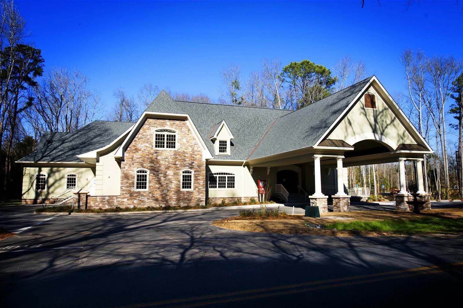 A large white house with a gray roof is surrounded by trees on a sunny day.