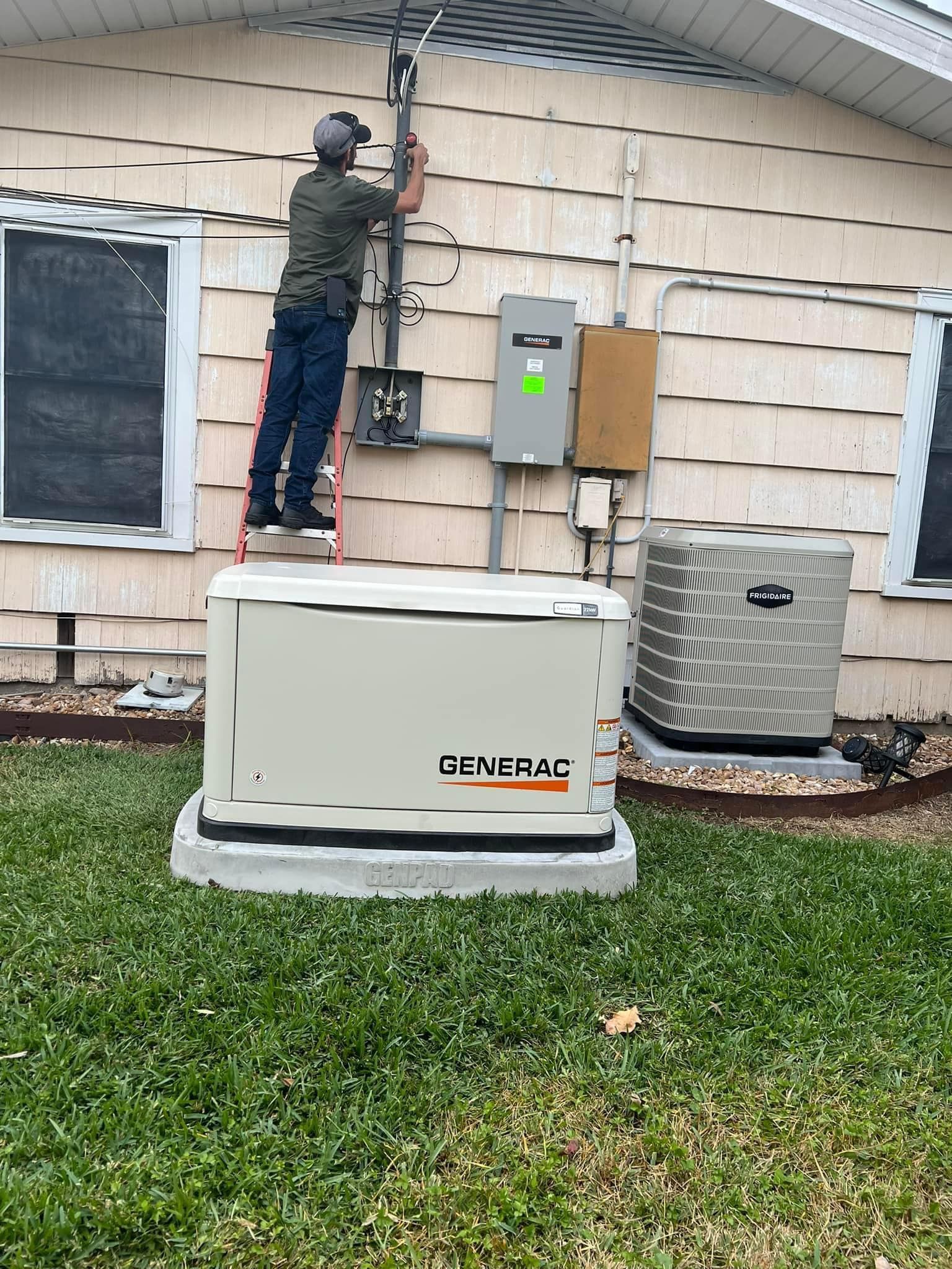 A man is standing on a ladder working on a generator outside of a house.