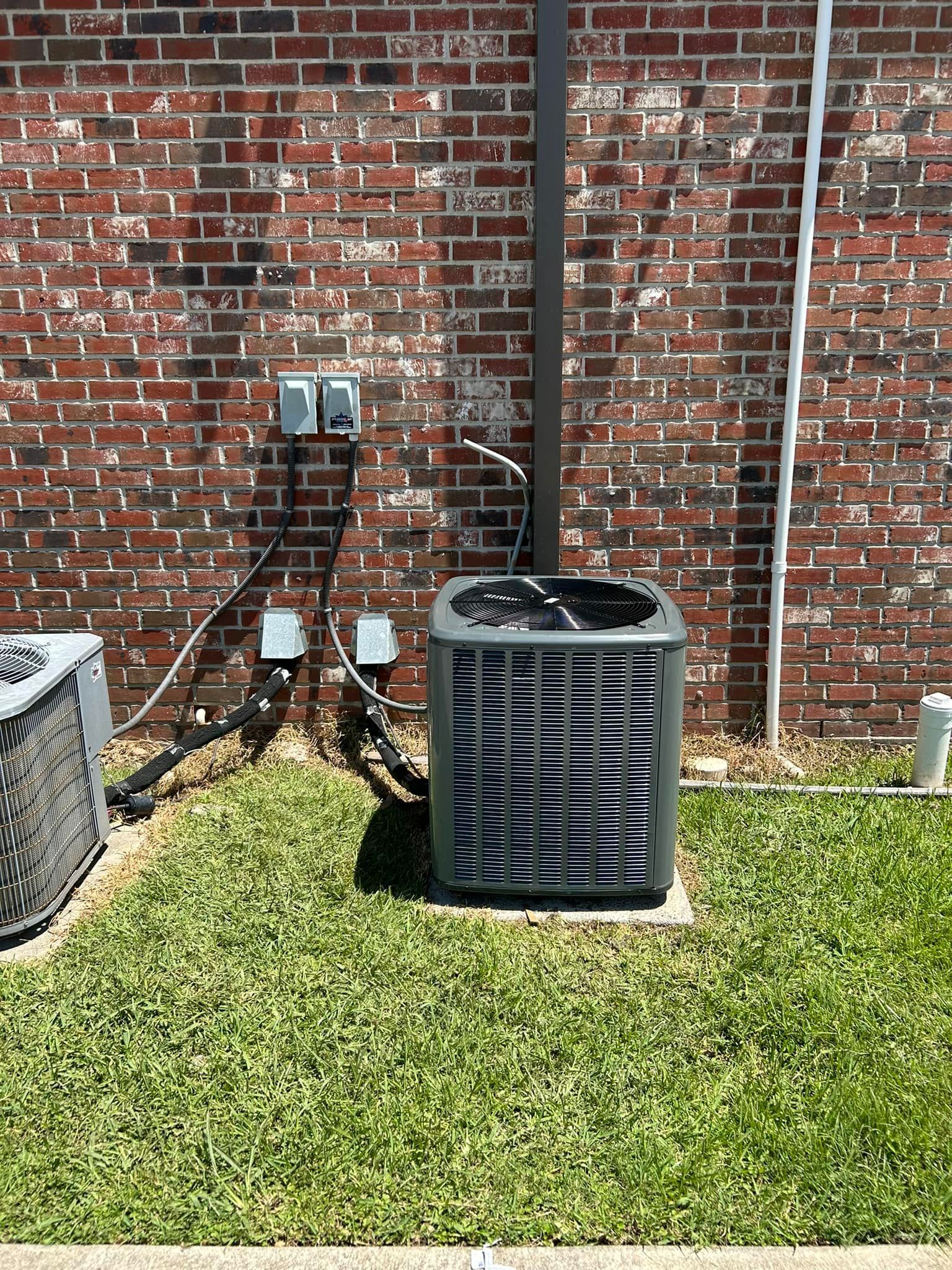 An air conditioner is sitting in the grass in front of a brick wall.