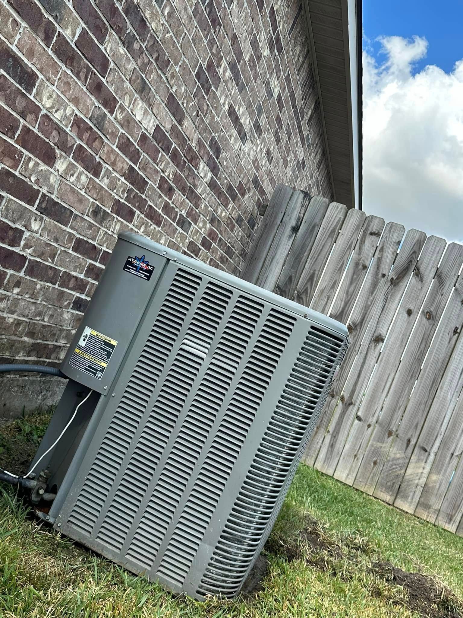 An air conditioner is sitting on the side of a brick building next to a wooden fence.