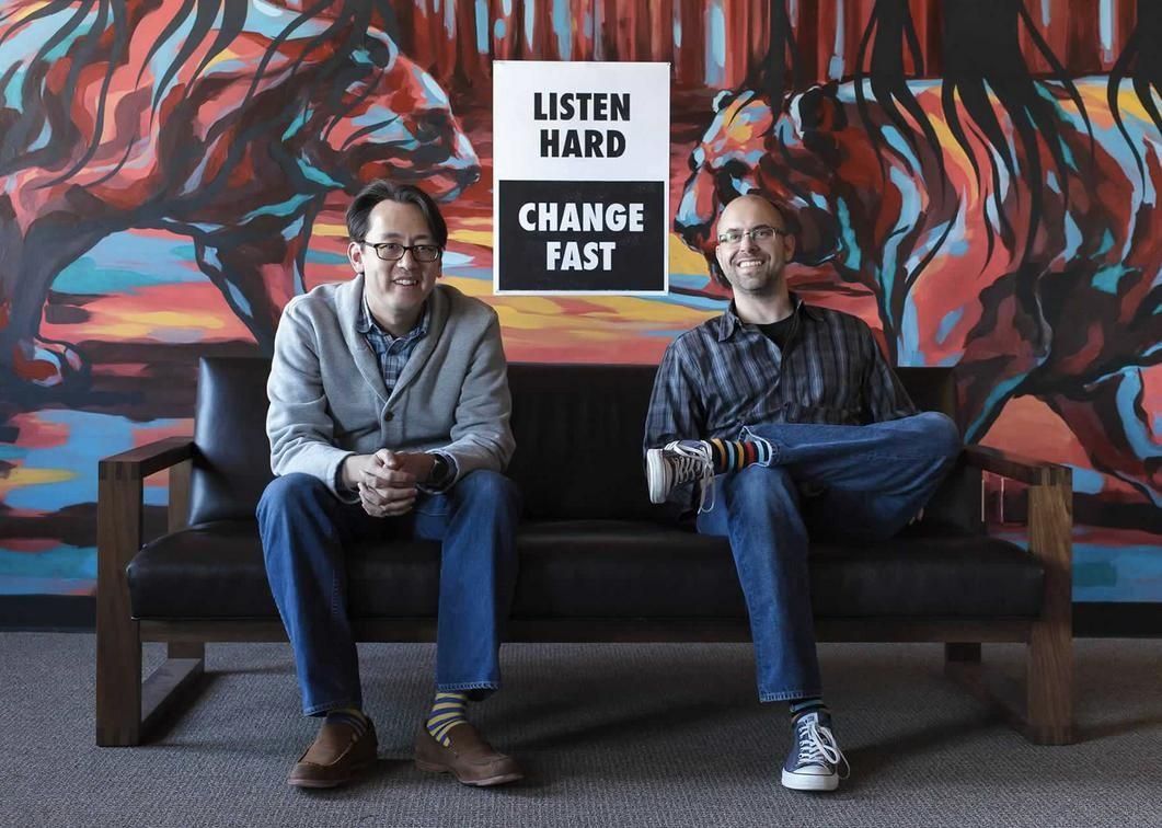 Two men sit on a couch with a sign that says listen hard change fast.
