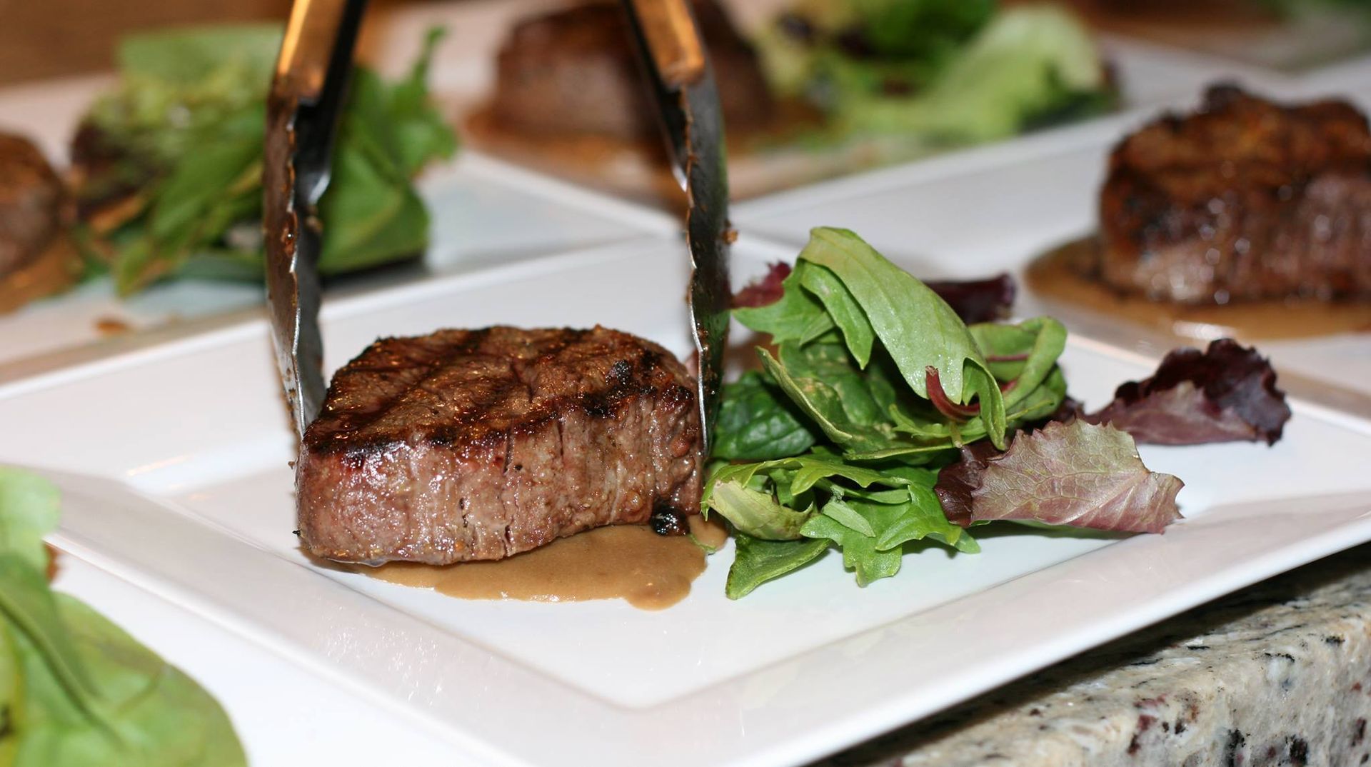 A steak is being served on a white plate with tongs.