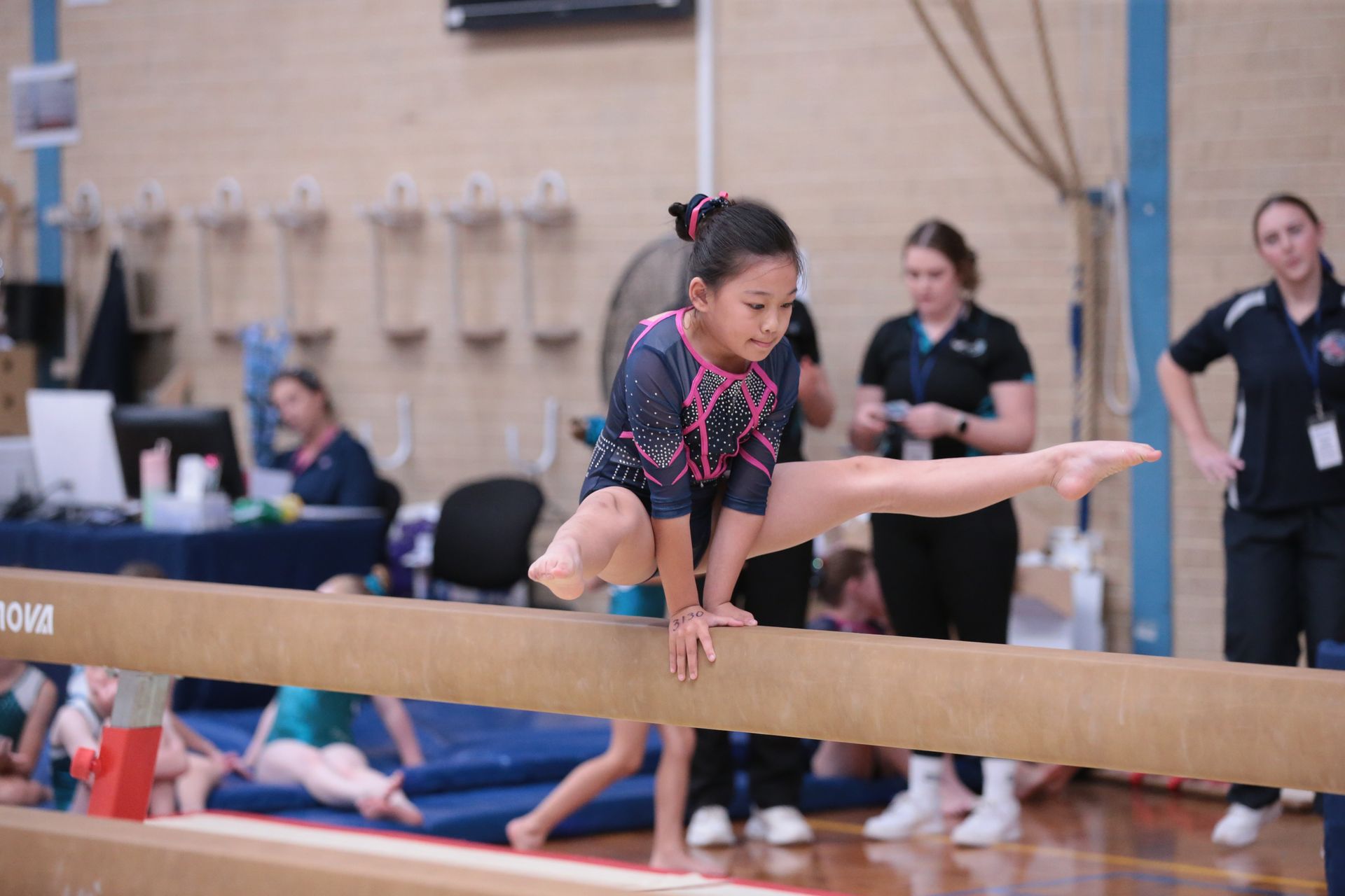 Gymnast on balance beam, performing a split. Indoor gym setting, judges watch.