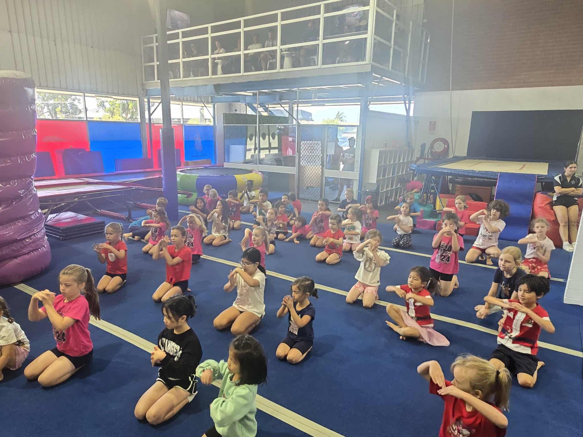 Children in a gym, kneeling and practicing dance moves. Many are in red. Blue mat, elevated area.