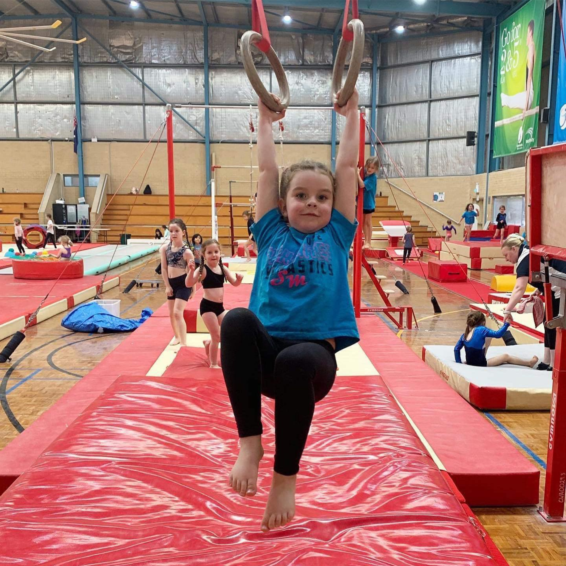 Girl hanging from gymnastic rings in gym. Blue shirt, black leggings, red mats.