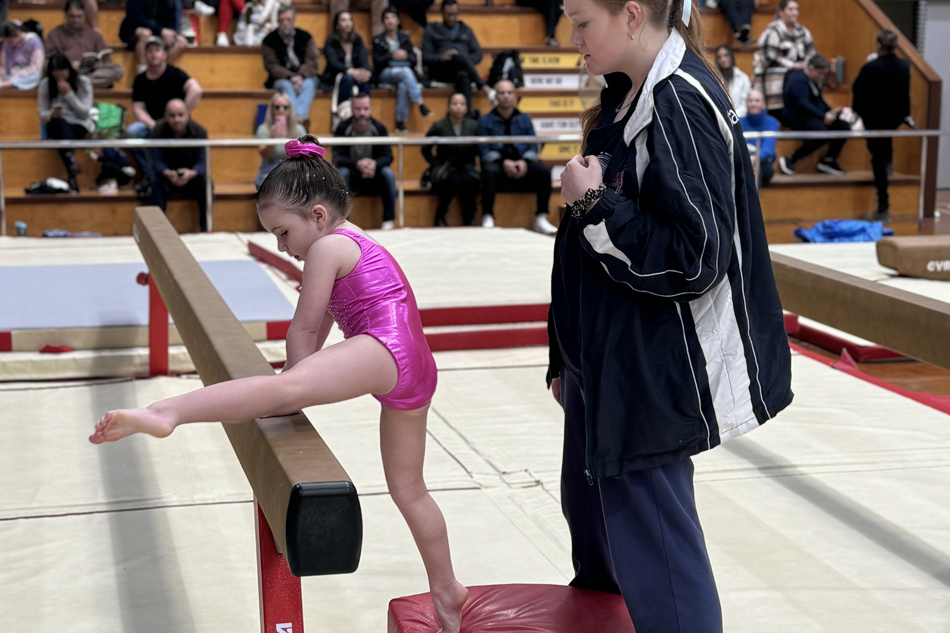 Young gymnast in pink leotard stretching on a balance beam, coach assisting in gym.