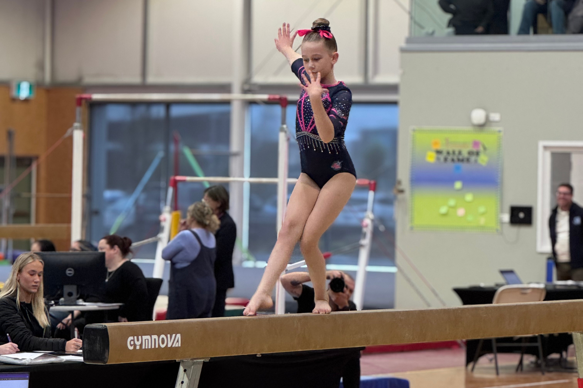 Young gymnast in a dark leotard balances on a beam, arms raised, in a gymnasium.