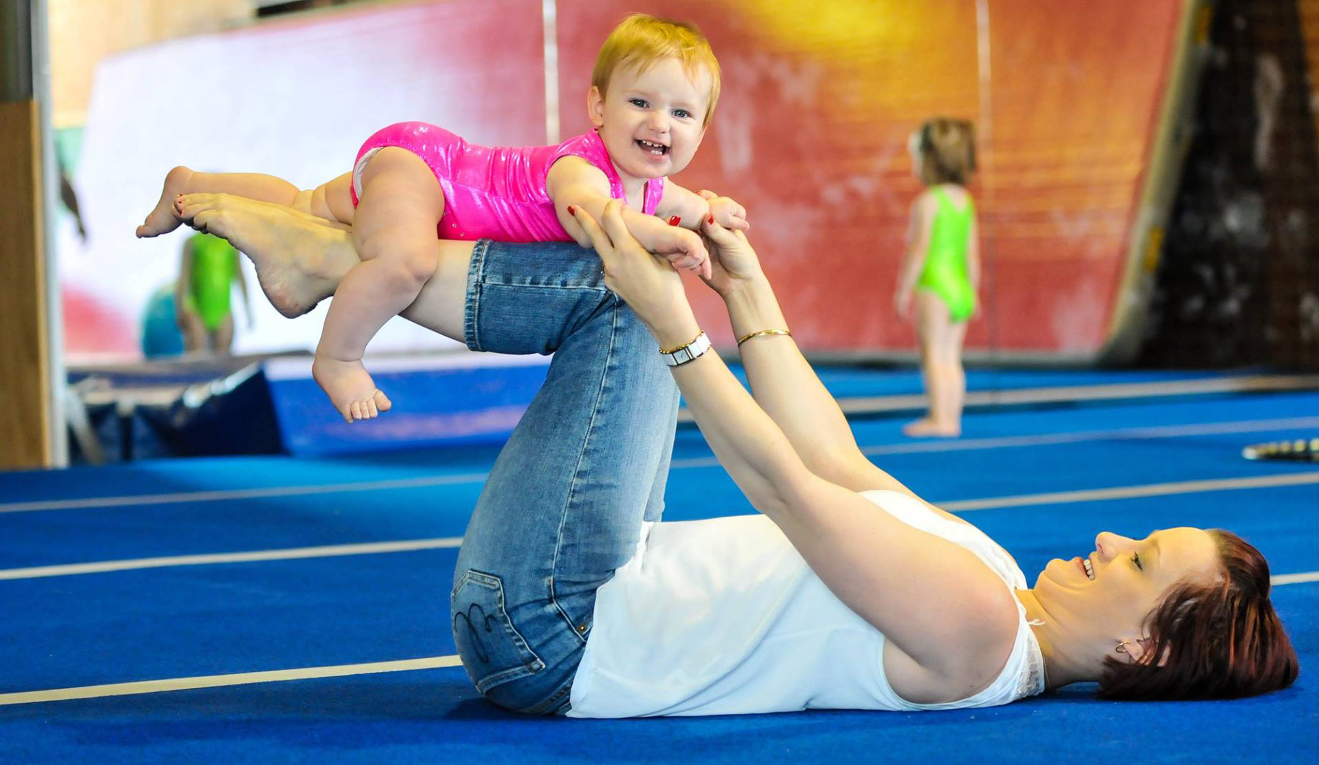 Woman lying on back supports baby in gymnastics pose; baby smiles. Blue mat.