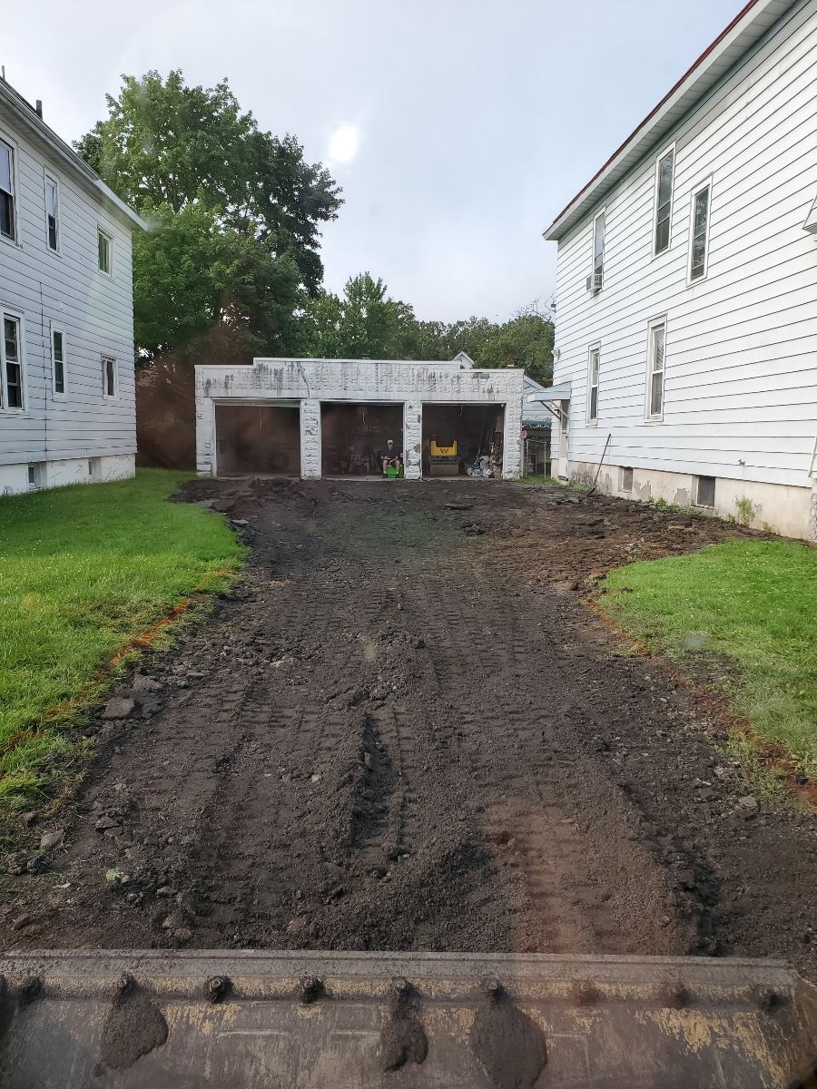 A tractor is driving down a dirt road in front of a house.