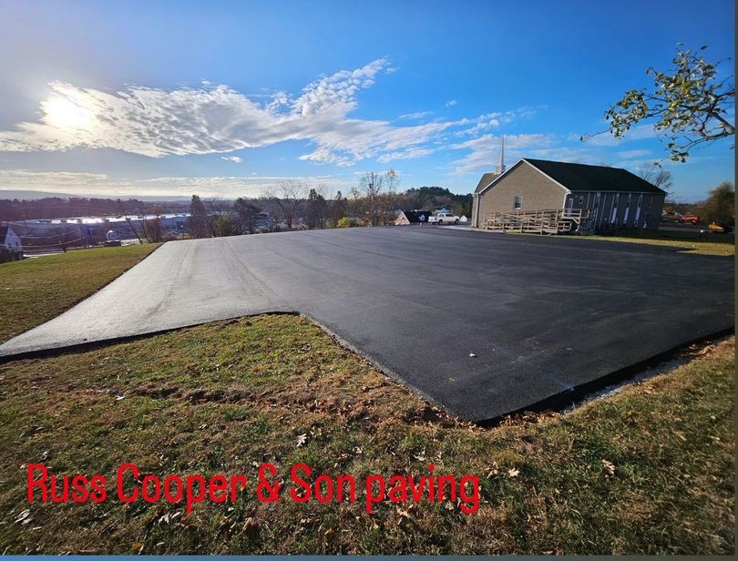 Newly paved asphalt parking area on a grassy hill, with a building in the background. Bright sky with clouds.