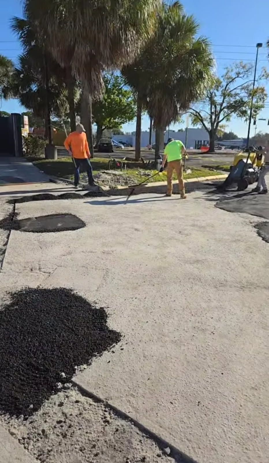 Road workers paving a street, asphalt spread on the pavement. Trees and buildings in the background. Sunny day.