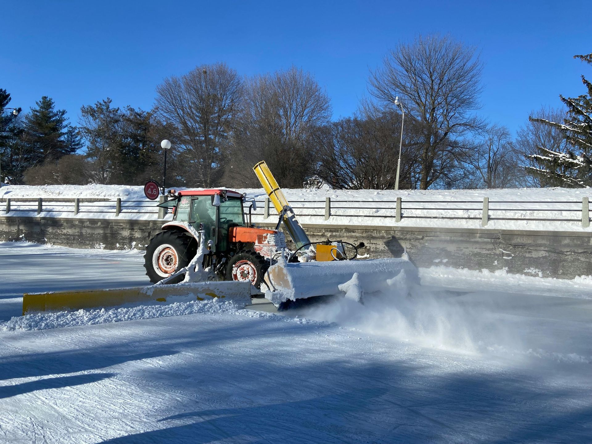 Tractor with snow blower clearing ice rink on a sunny day.
