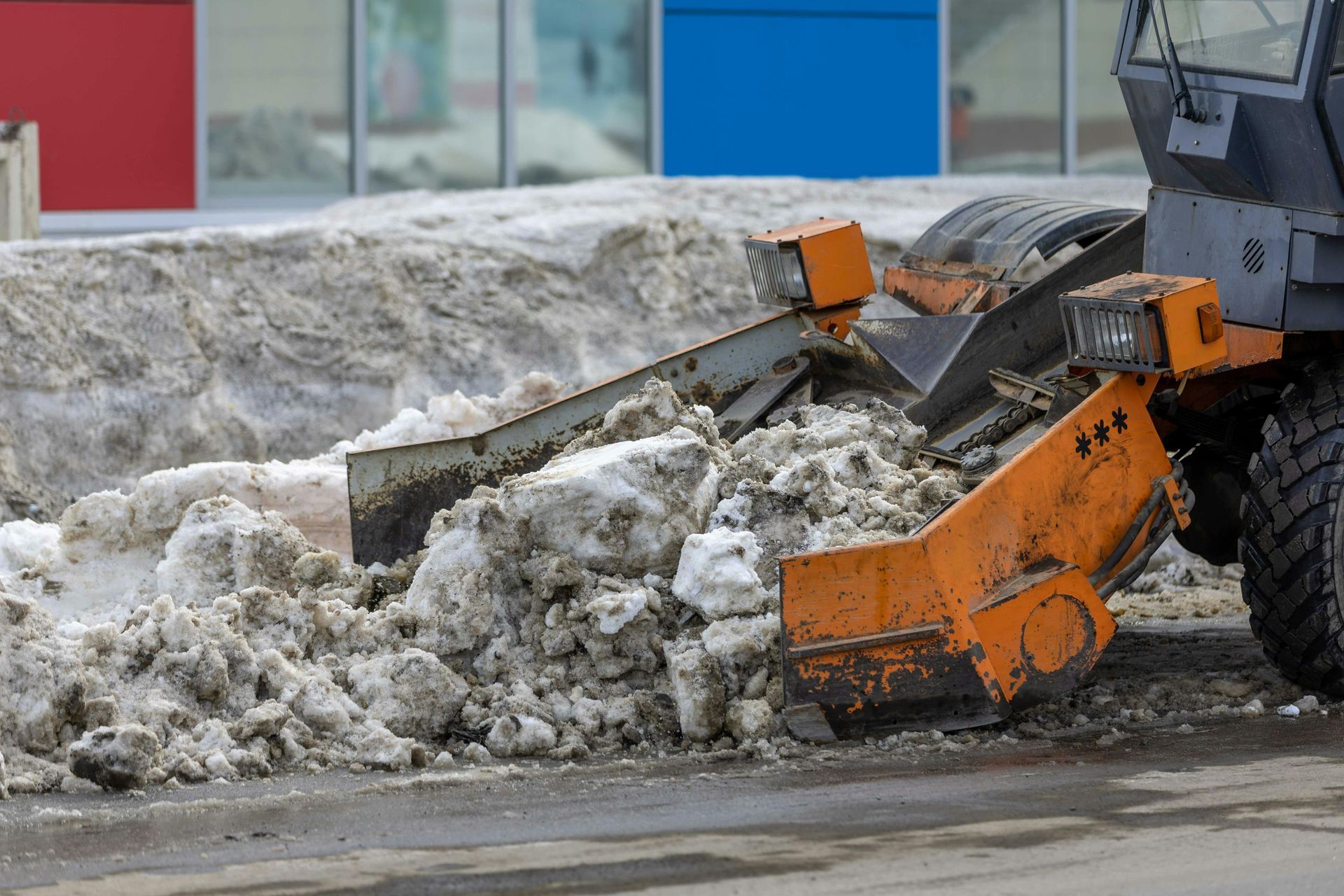 Orange snow blower pushing pile of dirty snow on pavement, blue and red building in background.